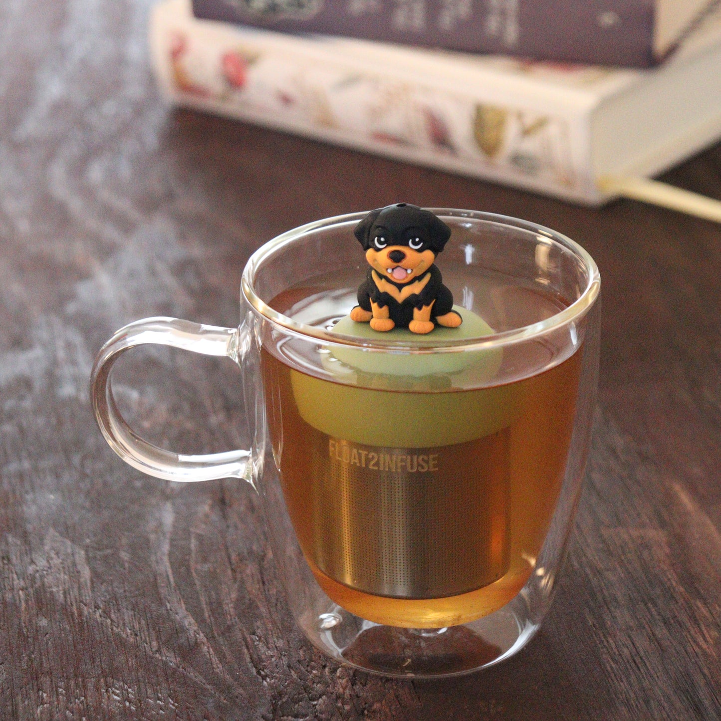 black and tan rottweiler dog animal tea infuser for loose leaf tea on a green base with a fine mesh stainless steel tea infuser that reads FLOAT2INFUSE floating in a glass tea mug with books in the background