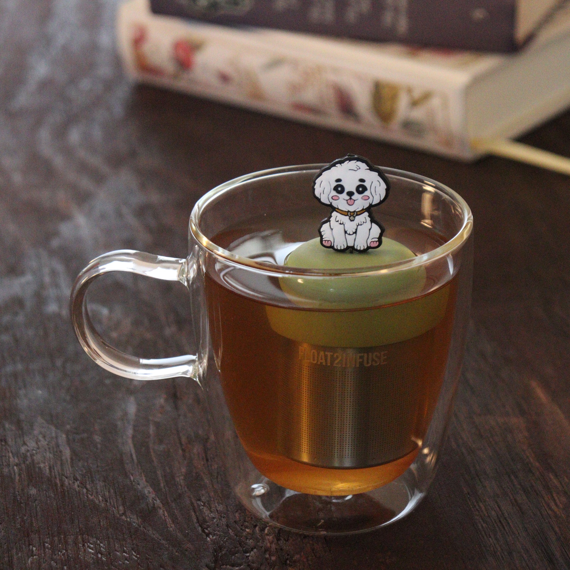 white maltipoo dog animal tea infuser for loose leaf tea on a green base with a fine mesh stainless steel tea infuser that reads FLOAT2INFUSE floating in a glass tea mug with books in the background