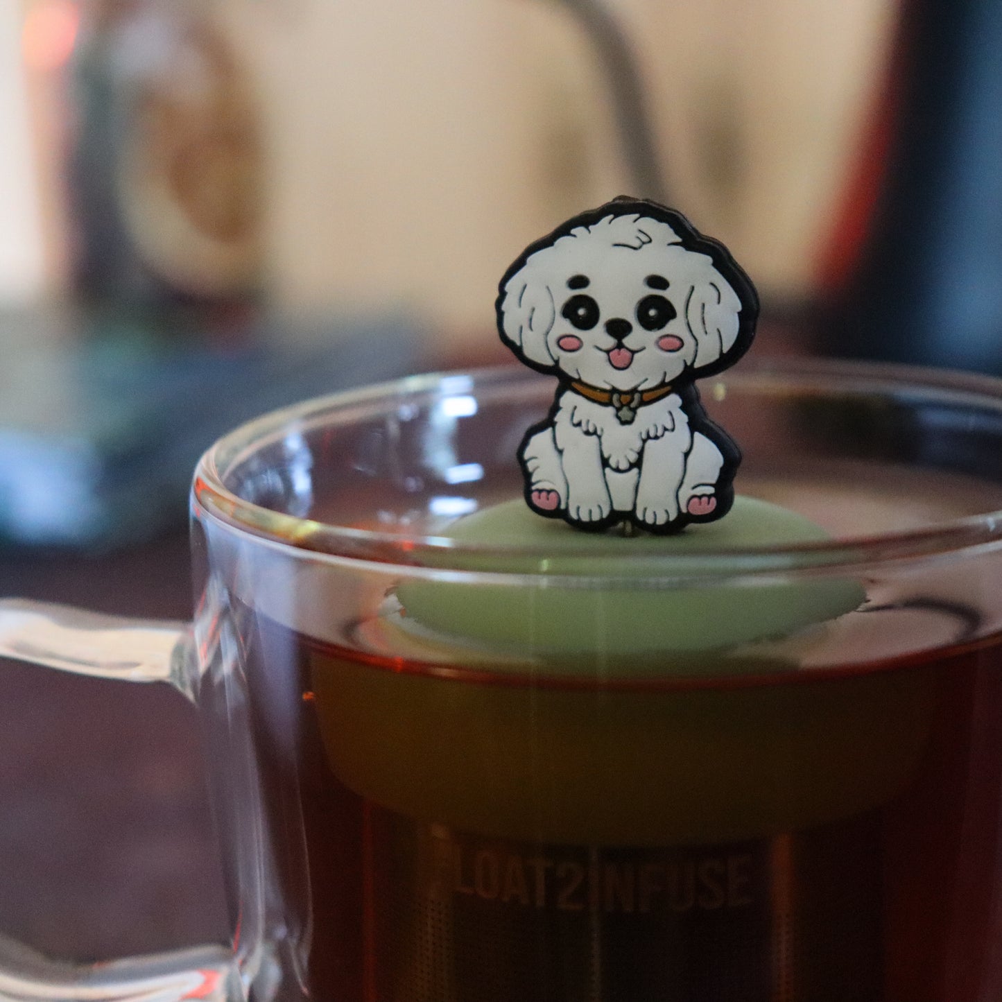 white maltipoo dog animal tea infuser for loose leaf tea on a green base with a fine mesh stainless steel tea strainer that reads FLOAT2INFUSE floating in a glass tea mug with tea kettle in the background