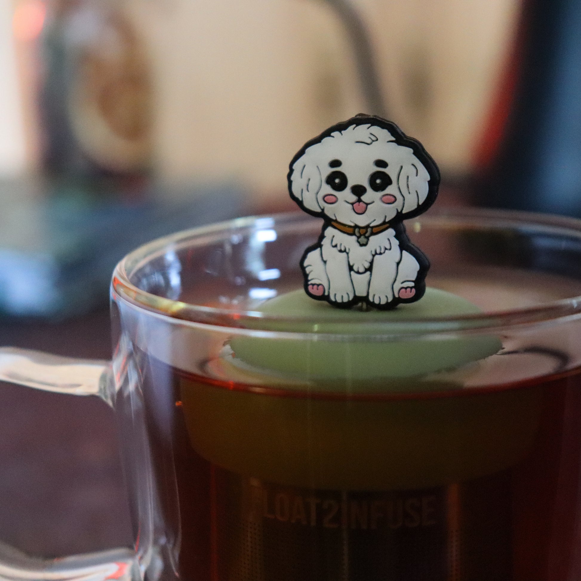 white maltipoo dog animal tea infuser for loose leaf tea on a green base with a fine mesh stainless steel tea strainer that reads FLOAT2INFUSE floating in a glass tea mug with tea kettle in the background