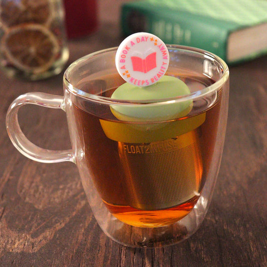 white circle that reads "a book a day keeps the reality away" with a centered pink book for loose leaf tea with a fine mesh stainless steel tea infuser that reads FLOAT2INFUSE on a green base  floating in a glass tea mug with  loose leaf tea and books in the background