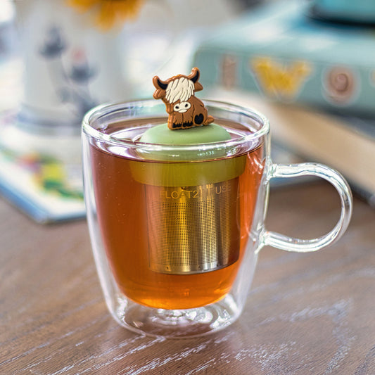 adult brown highland cow animal tea infuser for loose leaf tea with a fine mesh stainless steel tea strainer that reads FLOAT2INFUSE on a green base  floating in a glass tea mug with books in the background