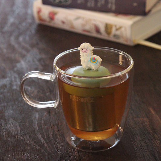 smiling alpaca animal tea infuser for loose leaf tea on a green base with a fine mesh stainless steel tea infuser that reads FLOAT2INFUSE floating in a glass tea mug with books in the background