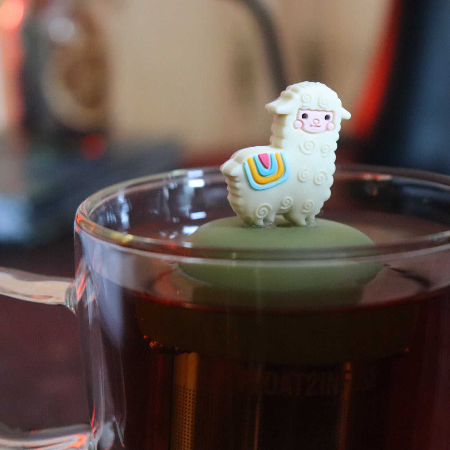 cute alpaca animal tea infuser for loose leaf tea with a fine mesh stainless steel tea strainer that reads FLOAT2INFUSE on a green base floating in a glass tea mug with tea kettle in the background