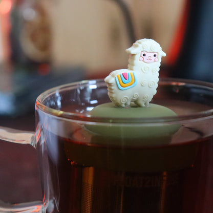 cute alpaca animal tea infuser for loose leaf tea with a fine mesh stainless steel tea strainer that reads FLOAT2INFUSE on a green base floating in a glass tea mug with tea kettle in the background