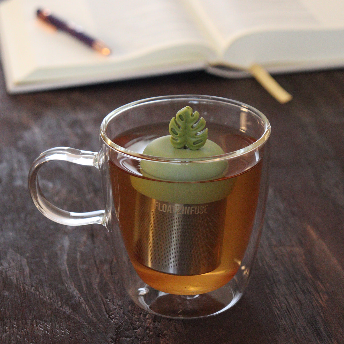 army green monstera leaf tea infuser for loose leaf tea with a fine mesh stainless steel tea infuser that reads FLOAT2INFUSE on a green base  floating in a glass tea mug with notebook and pen in the background