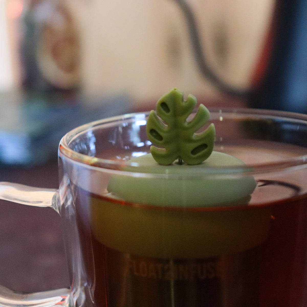 army green monstera leaf tea infuser for loose leaf tea  with a fine mesh stainless steel tea strainer that reads FLOAT2INFUSE on a green base floating in a glass tea mug with tea kettle in the background