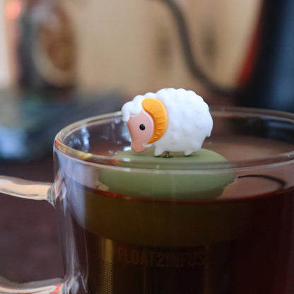 white baby bighorn sheep animal tea infuser for loose leaf tea  with a fine mesh stainless steel tea strainer that reads FLOAT2INFUSE on a green base floating in a glass tea mug with tea kettle in the background