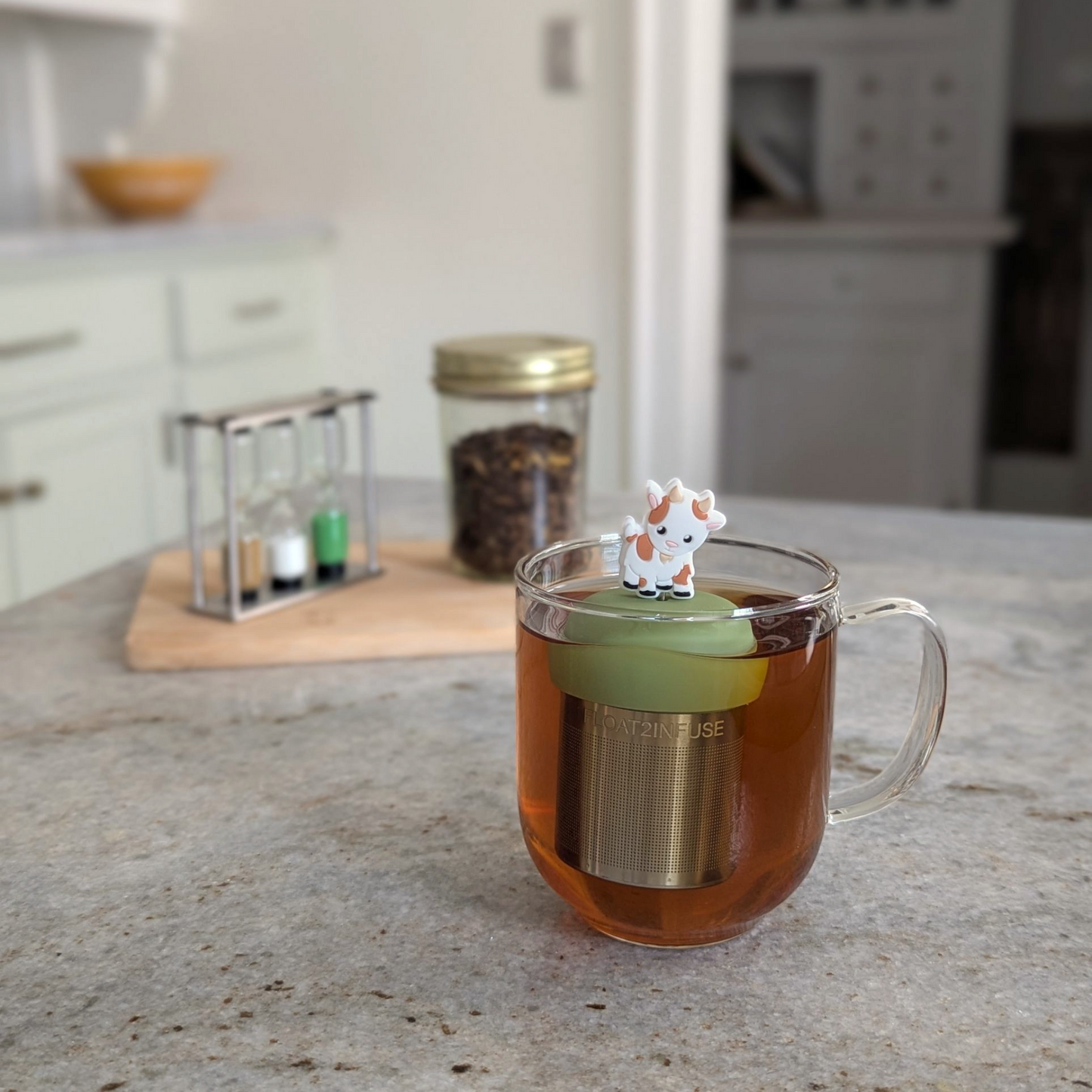 happy brown and white goat tea infuser for loose leaf tea  with a fine mesh stainless steel strainer that reads FLOAT2INFUSE on a green base floating in a glass tea mug with loose leaf tea in the background