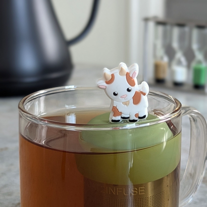 brown and white baby goat tea infuser for loose leaf tea  with a fine mesh stainless steel strainer that reads FLOAT2INFUSE on a green base floating in a glass tea mug with tea kettle in the background