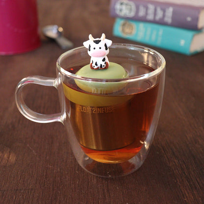 White with black spots baby cow infuser for loose leaf tea with a fine mesh stainless steel tea strainer 
that reads FLOAT2INFUSE on a green base with a tea canister and books in the background