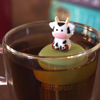 White with black spots baby cow infuser for loose leaf tea with a fine mesh stainless steel tea strainer 
that reads FLOAT2INFUSE on a green base with a tea canister and books in the background