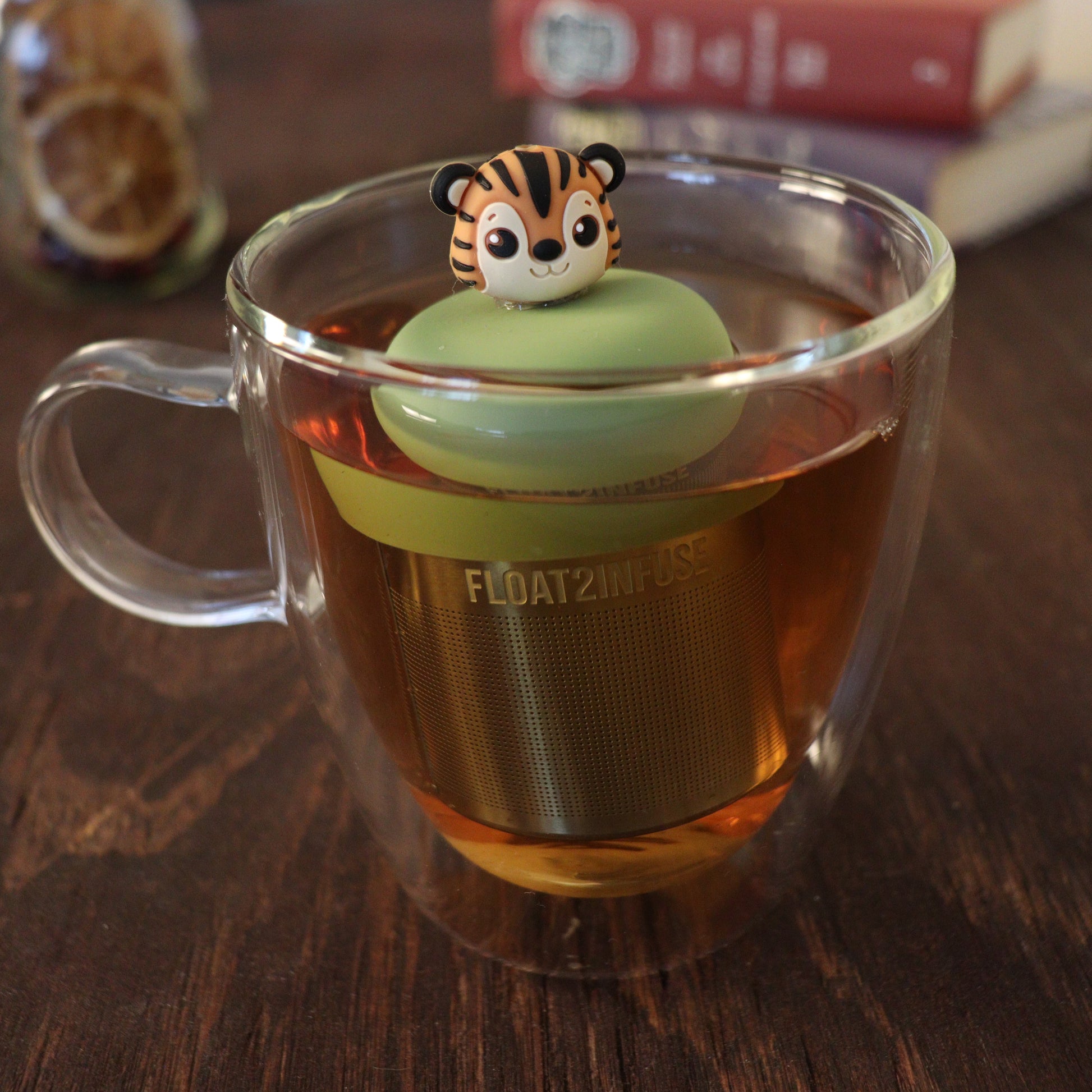 tiger tea infuser with a fine mesh stainless steel tea strainer that reads FLOAT2INFUSE with a green base floating in a glass tea mug with books in the background
