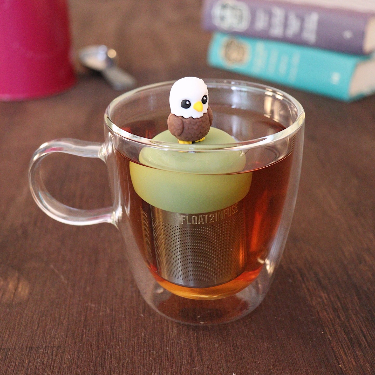 Cute bald eagle tea infuser for loose leaf tea with a fine mesh stainless steel tea strainer that reads FLOAT2INFUSE on a green base with a tea canister and books in the background