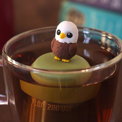 Cute bald eagle tea infuser for loose leaf tea with a fine mesh stainless steel tea strainer that reads FLOAT2INFUSE on a green base with a tea canister and books in the background