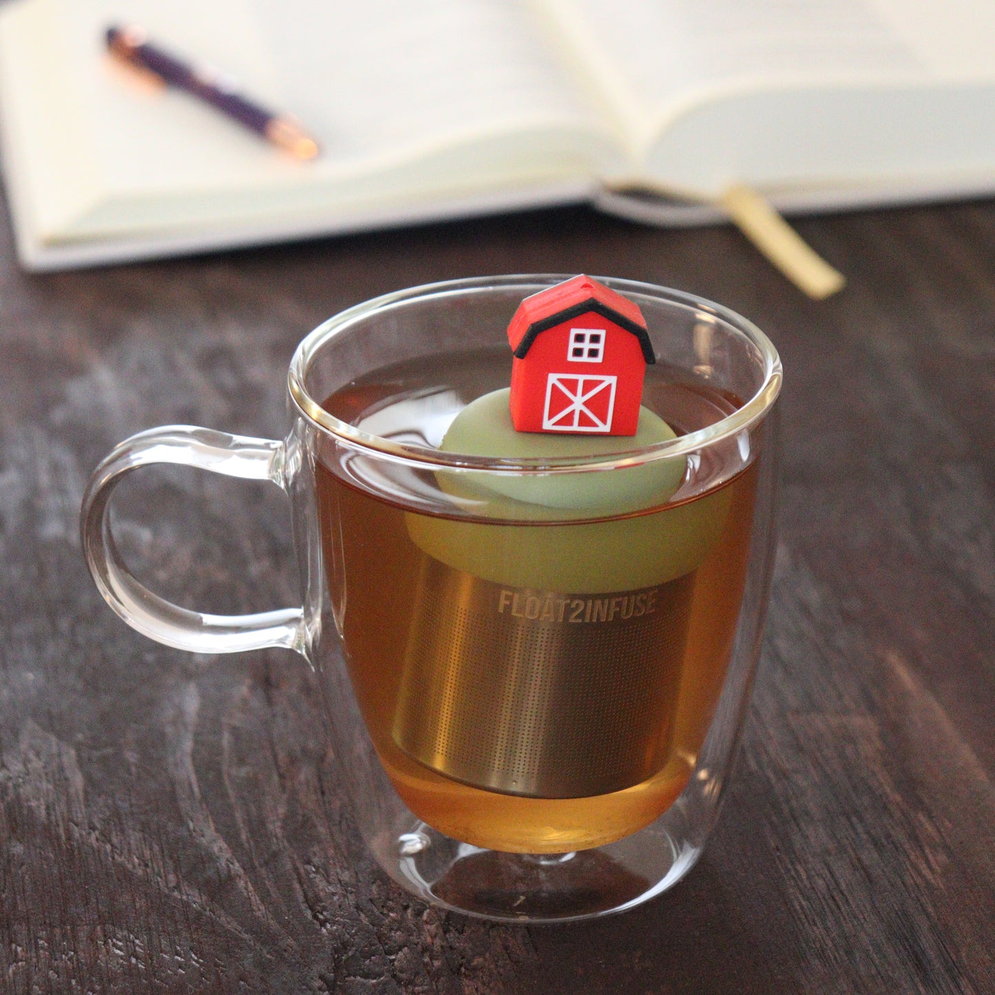 red barn tea infuser for loose leaf tea  with a fine mesh stainless steel tea infuser that reads FLOAT2INFUSE on a green base floating in a glass tea mug with notebook and pen in the background
