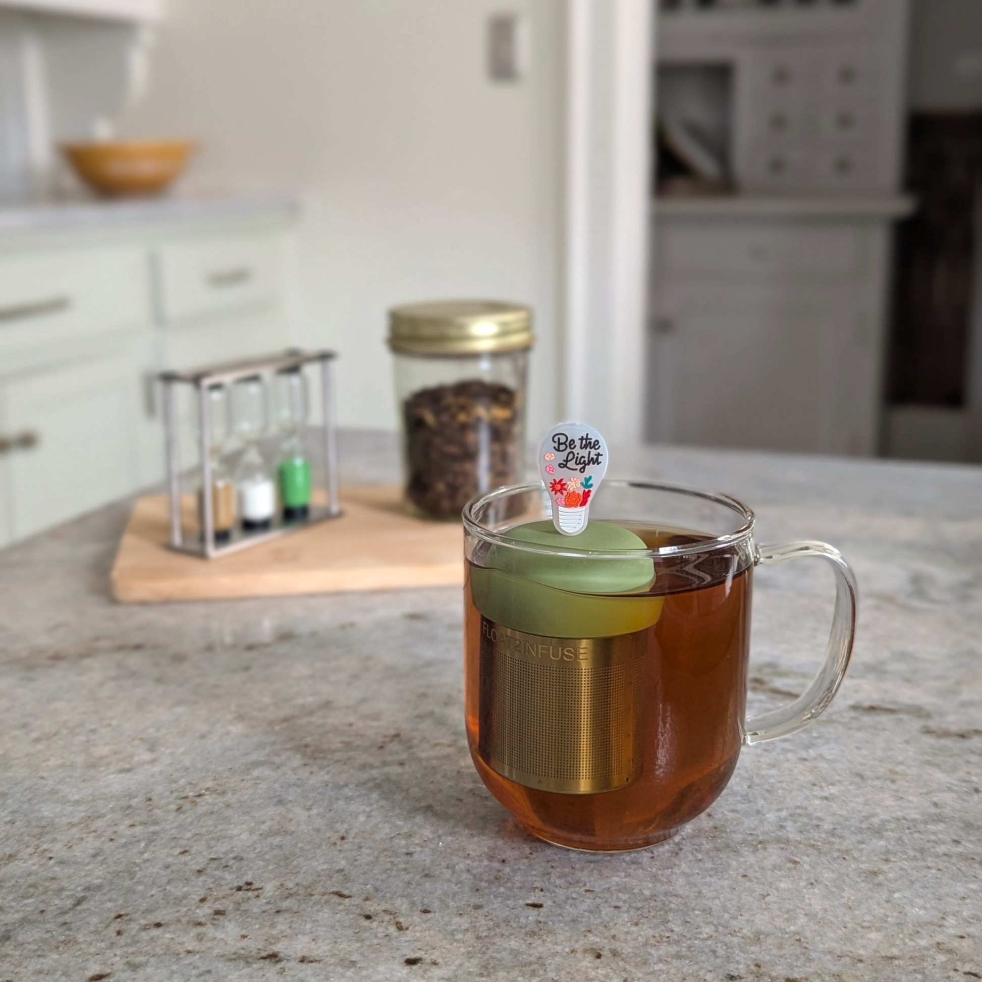 lightbulb with text "be the light" text and flower accents tea infuser for loose leaf tea  with a fine mesh stainless steel strainer that reads FLOAT2INFUSE on a green base floating in a glass tea mug with loose leaf tea in the background