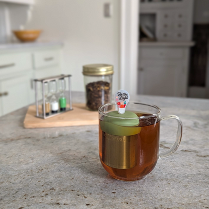 lightbulb with text "be the light" text and flower accents tea infuser for loose leaf tea  with a fine mesh stainless steel strainer that reads FLOAT2INFUSE on a green base floating in a glass tea mug with loose leaf tea in the background
