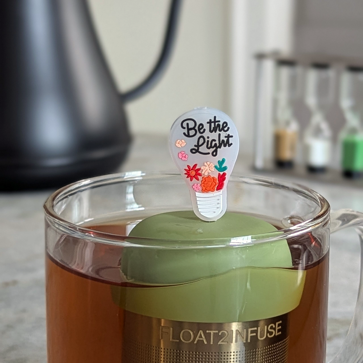 Lightbulb with the text 'Be the Light' message tea infuser for loose leaf tea  with a fine mesh stainless steel strainer that reads FLOAT2INFUSE on a green base floating in a glass tea mug with a tea kettle in the background