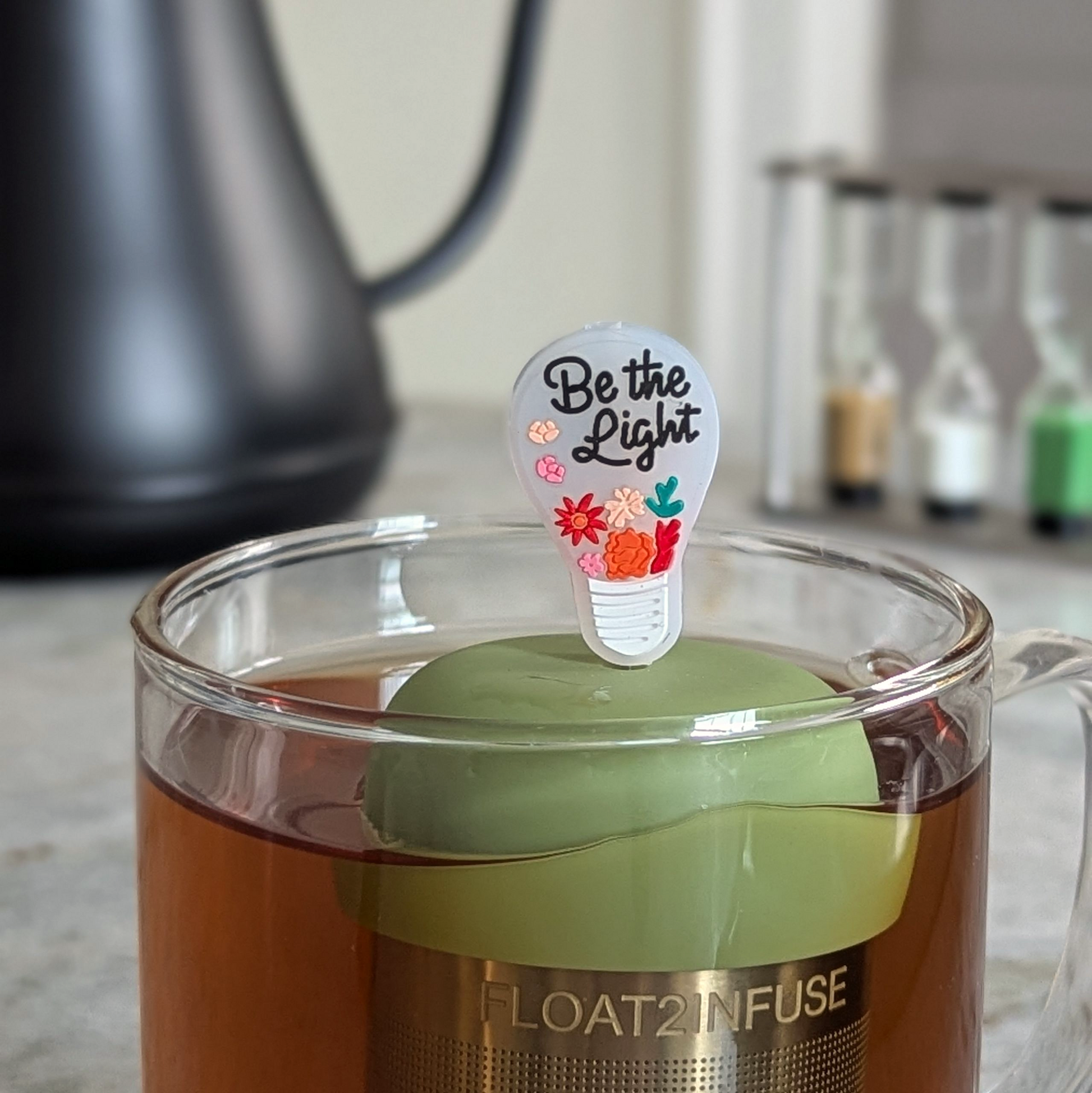 Lightbulb with the text 'Be the Light' message tea infuser for loose leaf tea  with a fine mesh stainless steel strainer that reads FLOAT2INFUSE on a green base floating in a glass tea mug with a tea kettle in the background