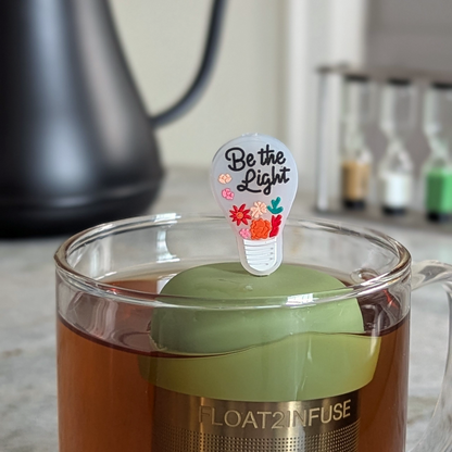 Lightbulb with the text 'Be the Light' message tea infuser for loose leaf tea  with a fine mesh stainless steel strainer that reads FLOAT2INFUSE on a green base floating in a glass tea mug with a tea kettle in the background