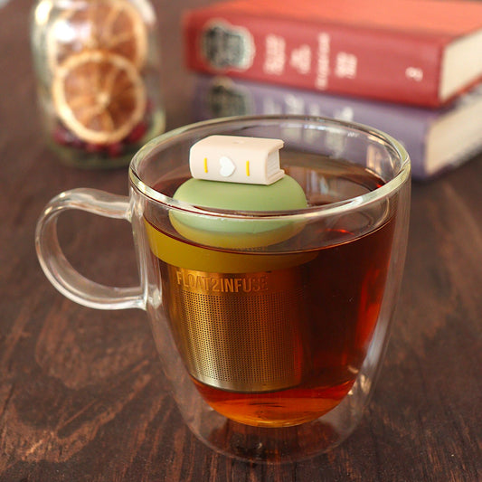 beige book with white heart tea infuser for loose leaf tea on a green base with a fine mesh stainless steel tea infuser that reads FLOAT2INFUSE floating in a glass tea mug with loose leaf tea and books in the background