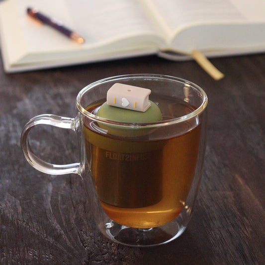 beige book with white heart tea infuser for loose leaf tea  with a fine mesh stainless steel tea infuser that reads FLOAT2INFUSE on a green base floating in a glass tea mug with notebook and pen in the background