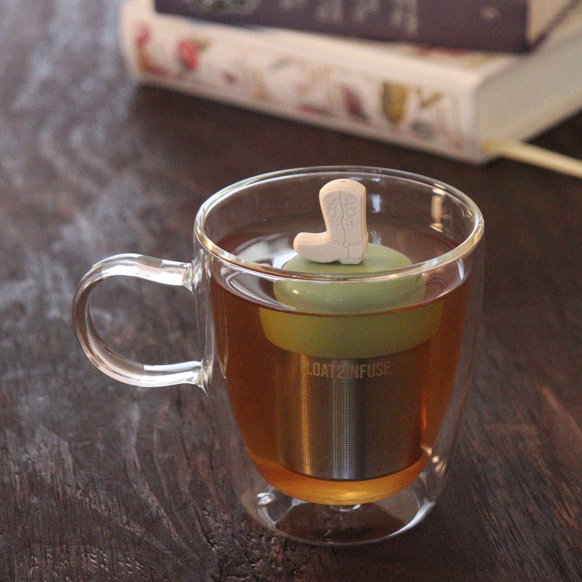 beige boot tea infuser for loose leaf tea on a green base with a fine mesh stainless steel tea infuser that reads FLOAT2INFUSE floating in a glass tea mug with books in the background