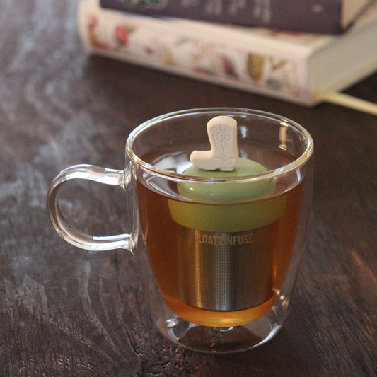 beige boot tea infuser for loose leaf tea on a green base with a fine mesh stainless steel tea infuser that reads FLOAT2INFUSE floating in a glass tea mug with books in the background