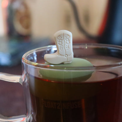 beige boot tea infuser for loose leaf tea  with a fine mesh stainless steel tea strainer that reads FLOAT2INFUSE on a green base floating in a glass tea mug with tea kettle in the background