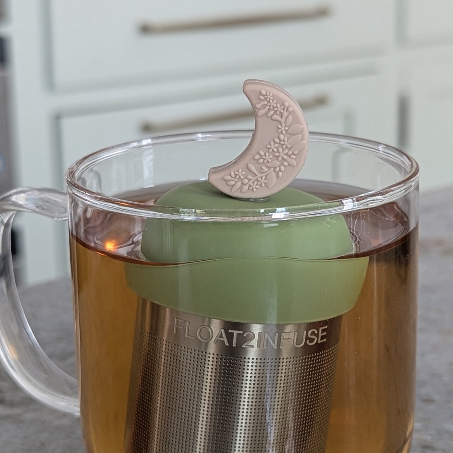 beige crescent moon with raised floral pattern tea infuser  for loose leaf tea with fine mesh stainless steel tea strainer that reads FLOAT2INFUSE on a green base floating in a glass tea mug