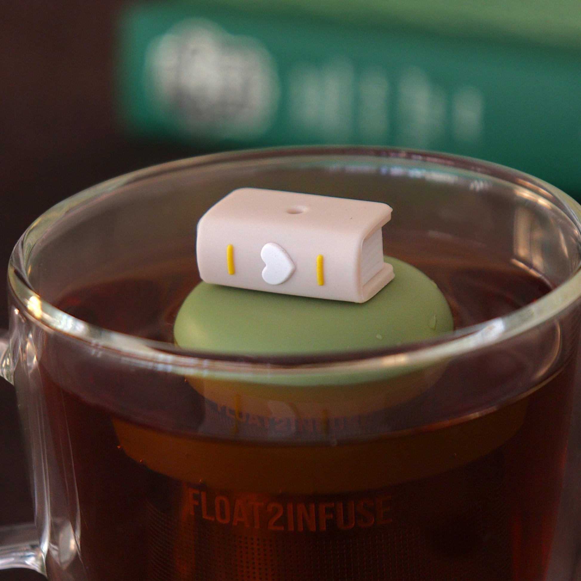 beige book with white heart tea infuser for loose leaf tea with a fine mesh stainless steel tea infuser that reads FLOAT2INFUSE on a green base  floating in a glass tea mug with books in the background