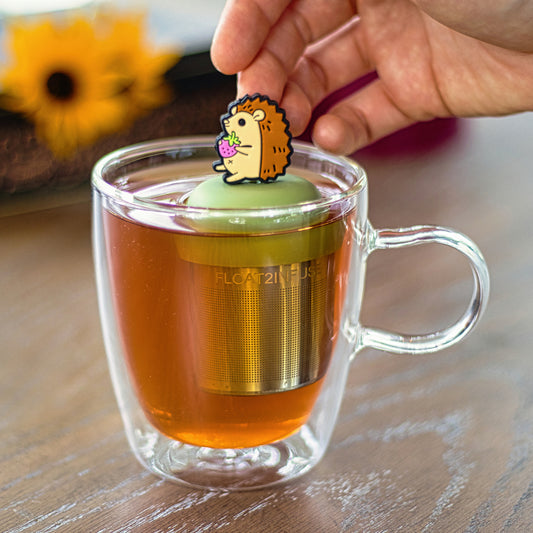 brown hedgehog holding a pink strawberry tea infuser for loose leaf tea  with a fine mesh stainless steel strainer that reads FLOAT2INFUSE on a green base floating in a glass tea mug with flowers in the background