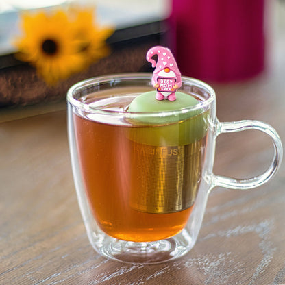 gnome with long white beard wearing pink clothing with white hearts holding a sign with the text "best mom ever" tea infuser for loose leaf tea with a fine mesh stainless steel tea strainer that reads FLOAT2INFUSE on a green base floating in a glass tea mug 
with flowers in the background