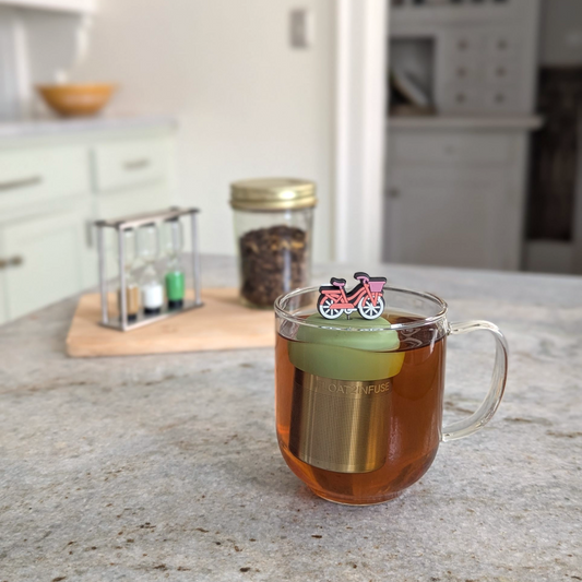 pink bicycle with white wheels and a purple basket tea infuser for loose leaf tea  with a fine mesh stainless steel strainer that reads FLOAT2INFUSE on a green base floating in a glass tea mug with loose leaf tea in the background