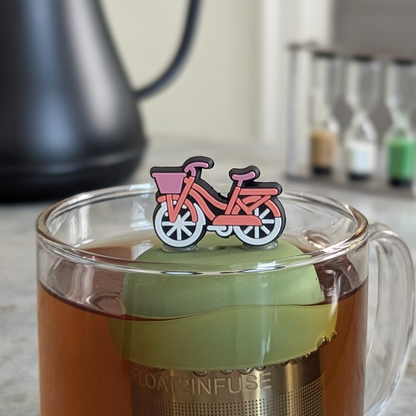 pink bicycle with white wheels and a purple basket tea infuser for loose leaf tea  with a fine mesh stainless steel strainer that reads FLOAT2INFUSE on a green base floating in a glass tea mug with a tea kettle in the background