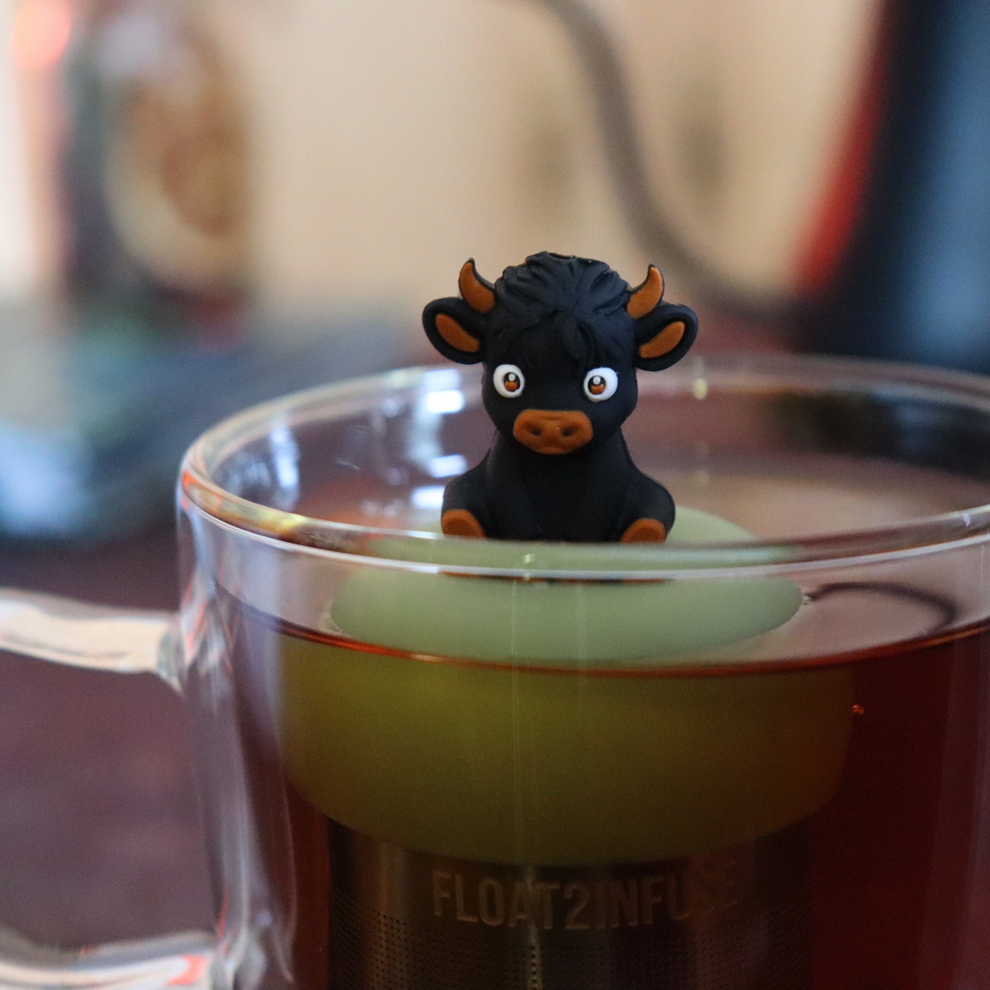 black baby highland cow animal tea infuser for loose leaf tea  with a fine mesh stainless steel tea strainer that reads FLOAT2INFUSE on a green base floating in a glass tea mug with tea kettle in the background