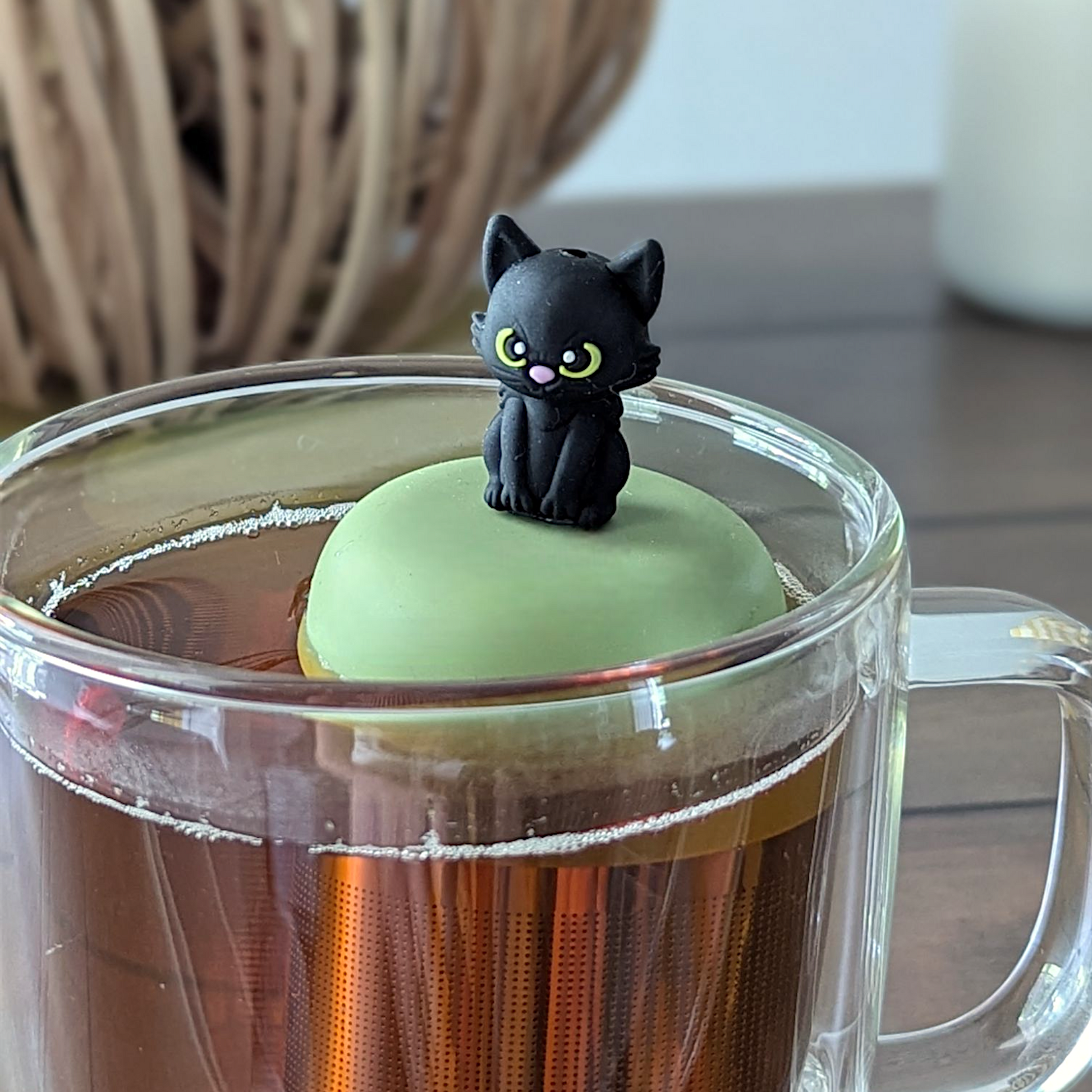 Black cat tea infuser for loose leaf tea with a fine mesh stainless steel tea strainer that reads FLOAT2INFUSE on a green base floating in a glass tea mug with a decorative pumpkin and white candle in the background