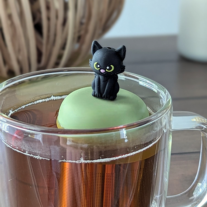 Black cat tea infuser for loose leaf tea with a fine mesh stainless steel tea strainer that reads FLOAT2INFUSE on a green base floating in a glass tea mug with a decorative pumpkin and white candle in the background