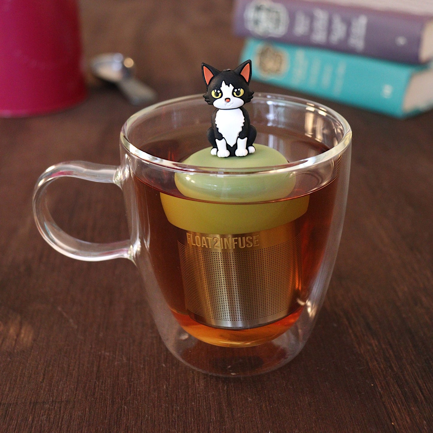 black and white cat tea infuser for loose leaf tea with a fine mesh stainless steel tea strainer 
that reads FLOAT2INFUSE on a green base with a tea canister and books in the background