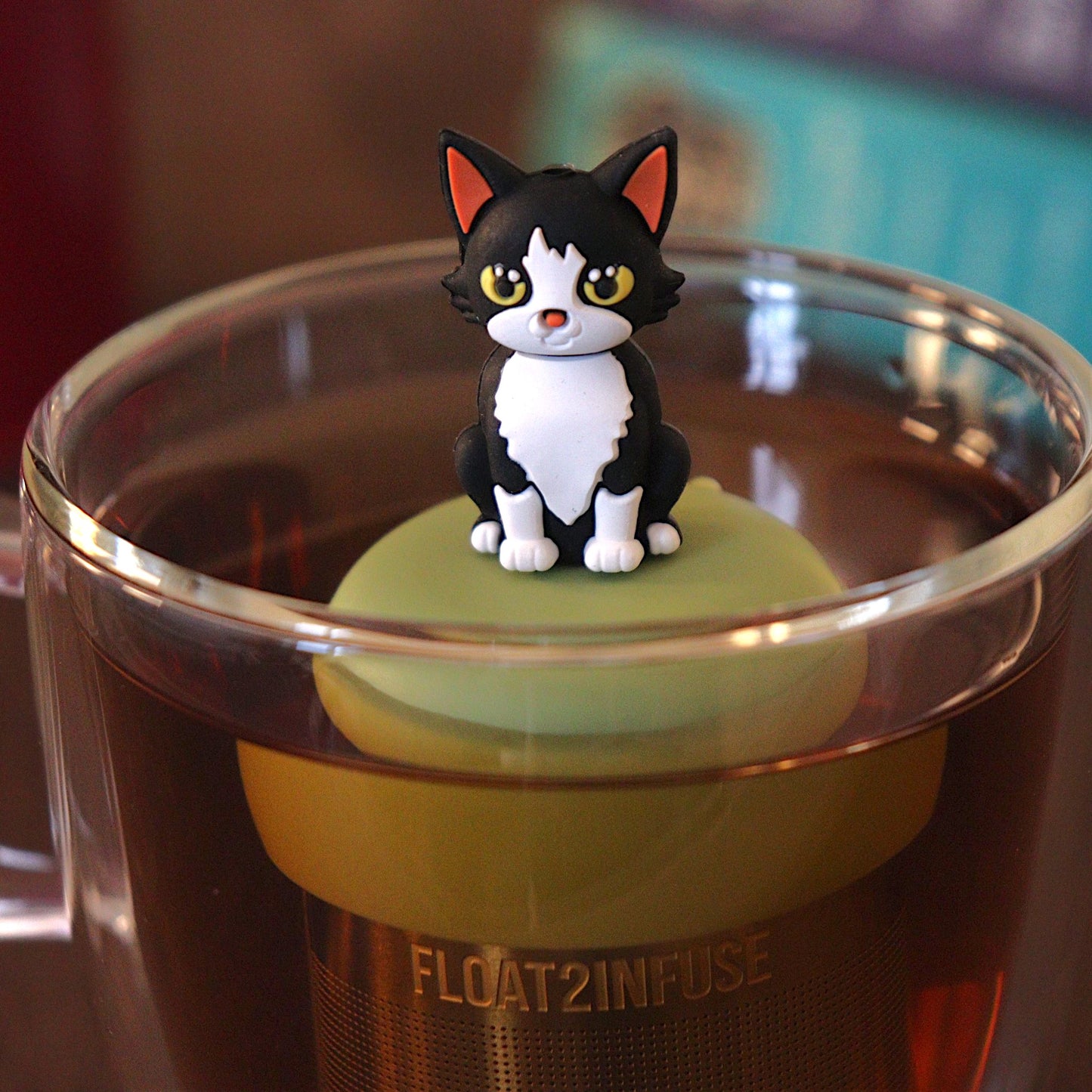 black and white cat tea infuser for loose leaf tea with a fine mesh stainless steel tea strainer 
that reads FLOAT2INFUSE on a green base with a tea canister and books in the background
