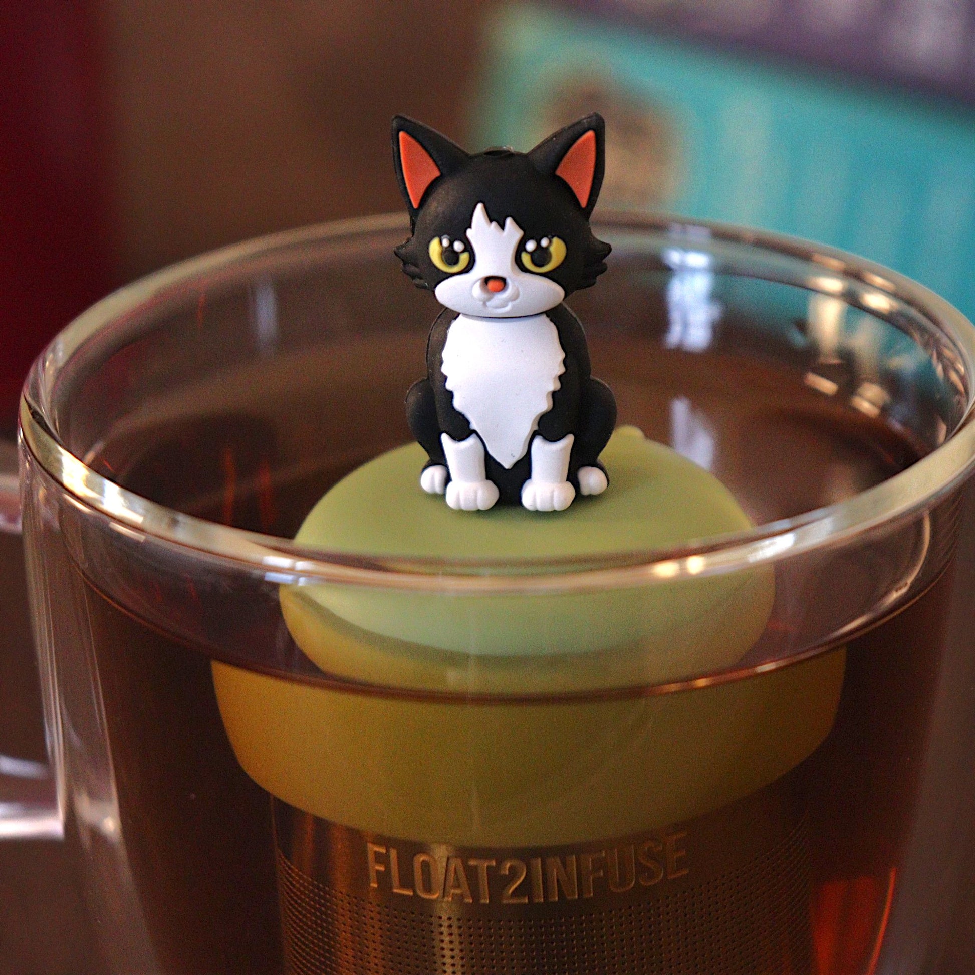 black and white cat tea infuser for loose leaf tea with a fine mesh stainless steel tea strainer 
that reads FLOAT2INFUSE on a green base with a tea canister and books in the background