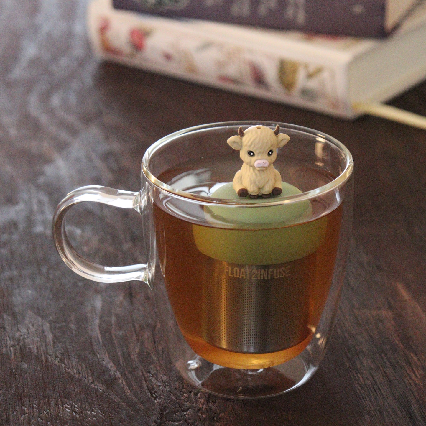 blonde baby highland cow animal tea infuser for loose leaf tea on a green base with a fine mesh stainless steel tea infuser that reads FLOAT2INFUSE floating in a glass tea mug with books in the background