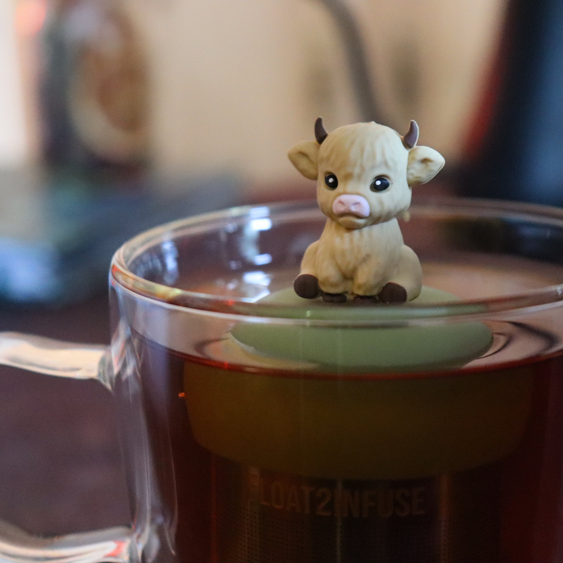 blonde baby cow animal tea infuser for loose leaf tea on a green base with a fine mesh stainless steel tea strainer that reads FLOAT2INFUSE floating in a glass tea mug with tea kettle in the background