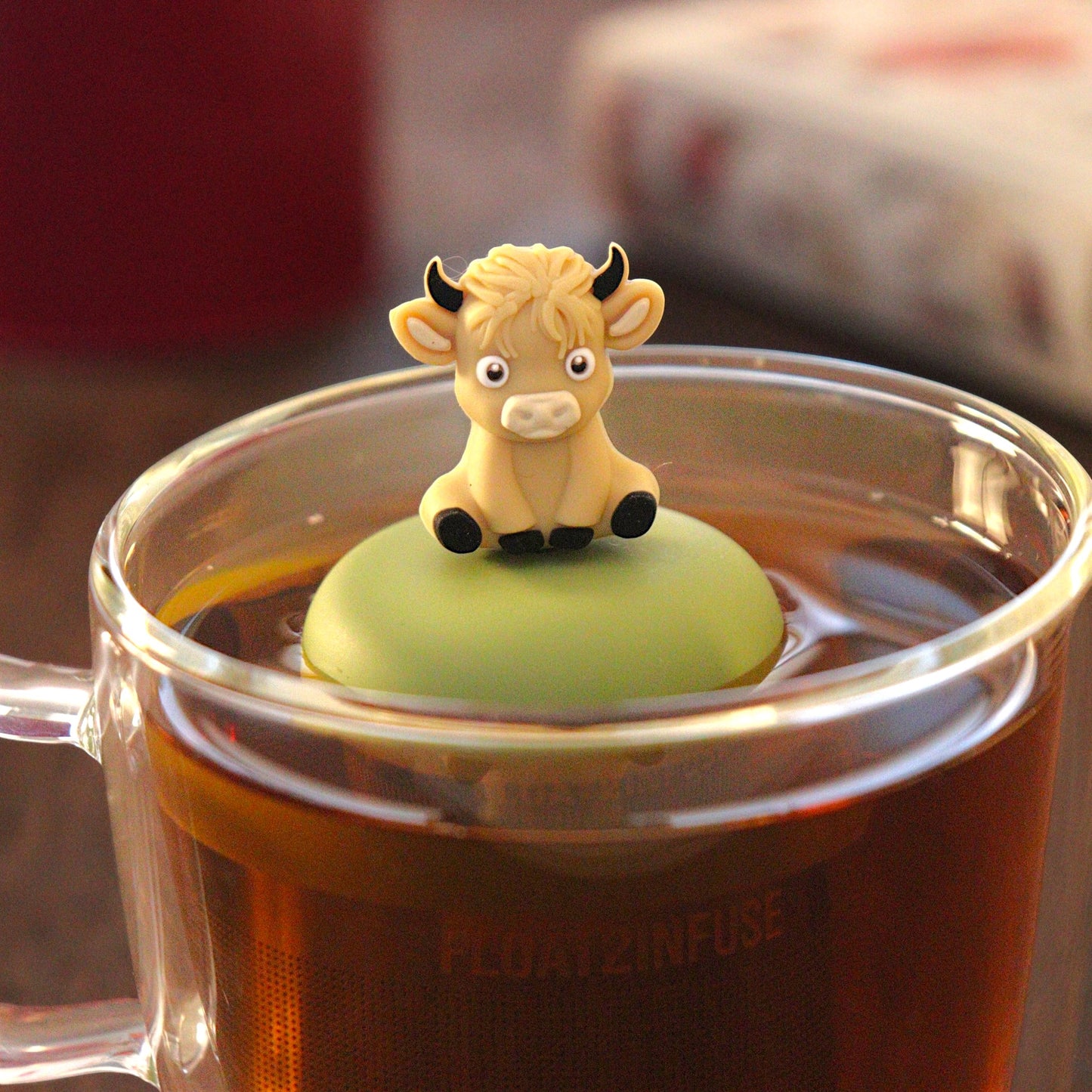 blonde baby highland cow animal tea infuser for loose leaf tea with a fine mesh stainless steel tea strainer that reads FLOAT2INFUSE on a green base   floating in a glass tea mug with tea kettle in the background