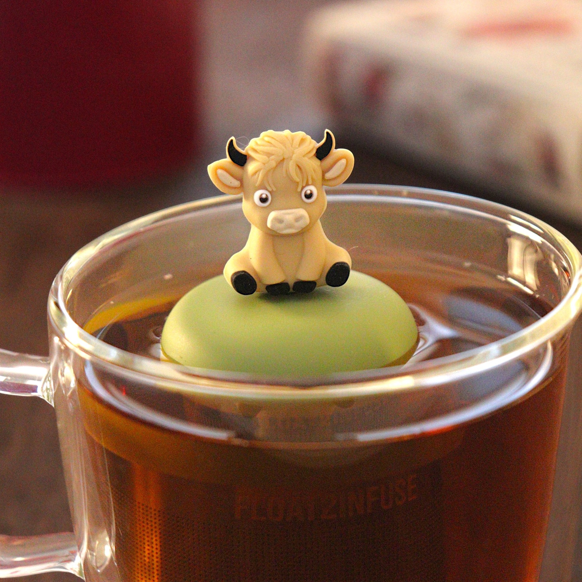 blonde baby highland cow animal tea infuser for loose leaf tea with a fine mesh stainless steel tea strainer that reads FLOAT2INFUSE on a green base   floating in a glass tea mug with tea kettle in the background
