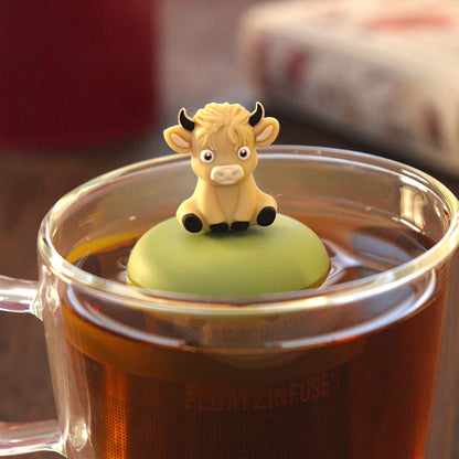 blonde baby highland cow animal tea infuser for loose leaf tea with a fine mesh stainless steel tea strainer that reads FLOAT2INFUSE on a green base   floating in a glass tea mug with tea kettle in the background