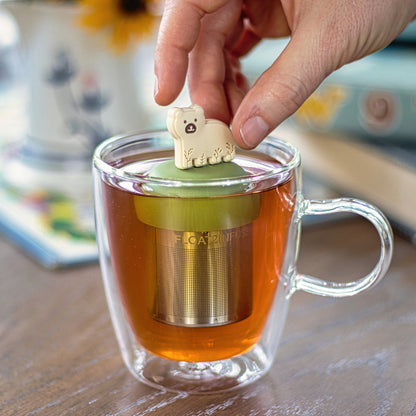 beige bear animal with leaf patterns  tea infuser for loose leaf tea with a fine mesh stainless steel tea strainer that reads FLOAT2INFUSE on a green base floating in a glass tea mug with books in the background
