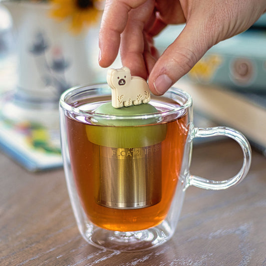 beige bear animal with leaf patterns  tea infuser for loose leaf tea with a fine mesh stainless steel tea strainer that reads FLOAT2INFUSE on a green base floating in a glass tea mug with books in the background