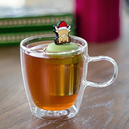 blonde highland cow animal wearing a santa hat tea infuser for loose leaf tea with a fine mesh stainless steel tea strainer that reads FLOAT2INFUSE on a green base  floating in a glass tea mug 
with books in the background
